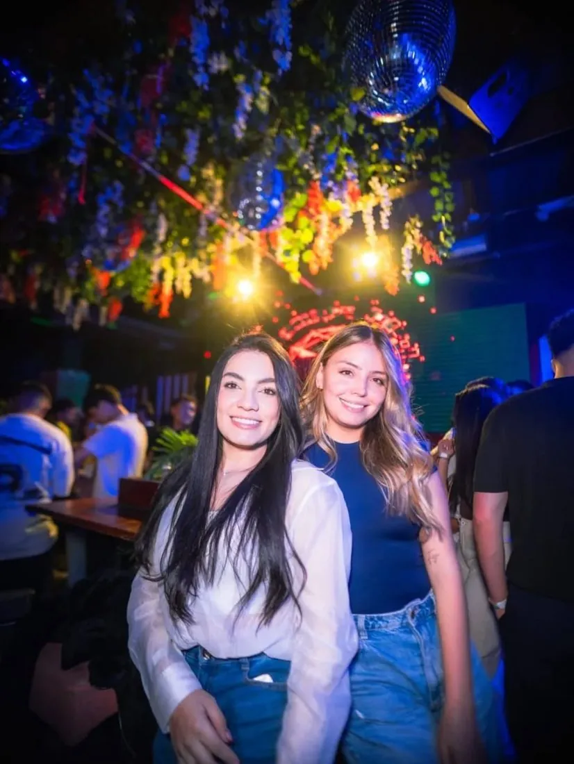 Dos amigas sonriendo en un bar con luces de colores y flores en el techo